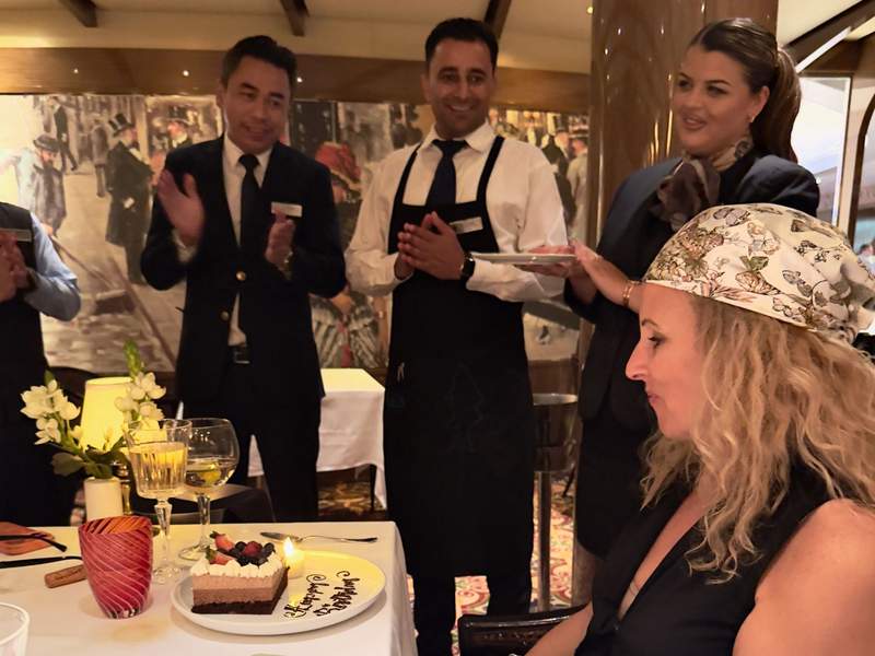 Servers clapping and presenting a birthday cake with a lit candle to a guest at a specialty restaurant table on Koningsdam.
