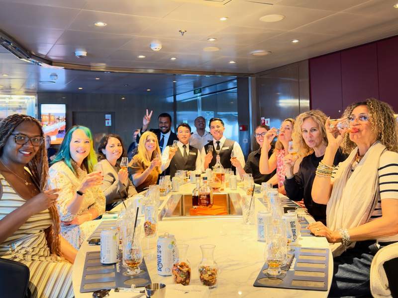 A diverse group of smiling women raise their tasting glasses during a guided Jefferson's Bourbon tasting class around a large marble bar on the Koningsdam.