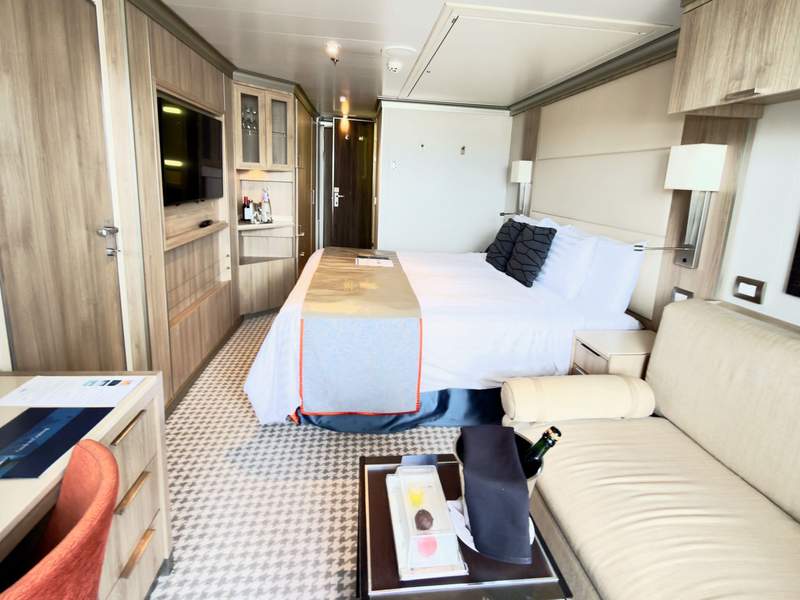 Cruise cabin with king bed covered by white duvet next to small couch and table with champagne in a bucket.