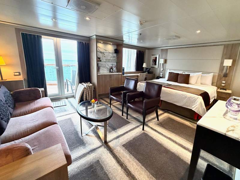King bed with leather chairs, table, full size pink velvet couch and balcony overlooking blue ocean on Holland America cruise ship.