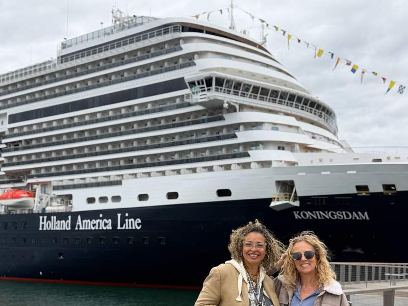 Two female friends smile and pose together on a wooden pier with the massive bow of the Holland America Line Koningsdam cruise ship behind them.