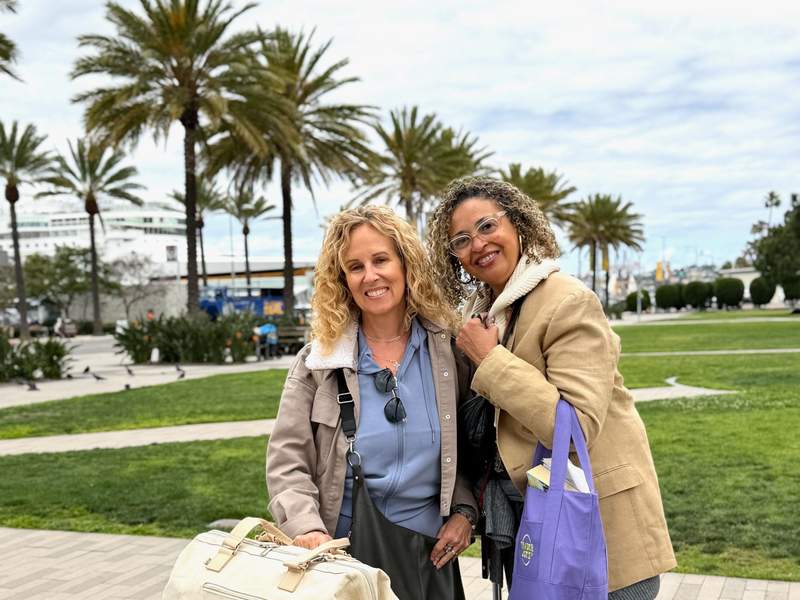 Two happy women smiling and holding bags at the San Diego port terminal with palm trees and a cruise ship in the background.