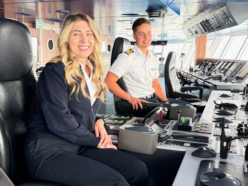 Blonde woman and man sitting in captains chairs with a dashboard of controls in front of them on a cruise ship.