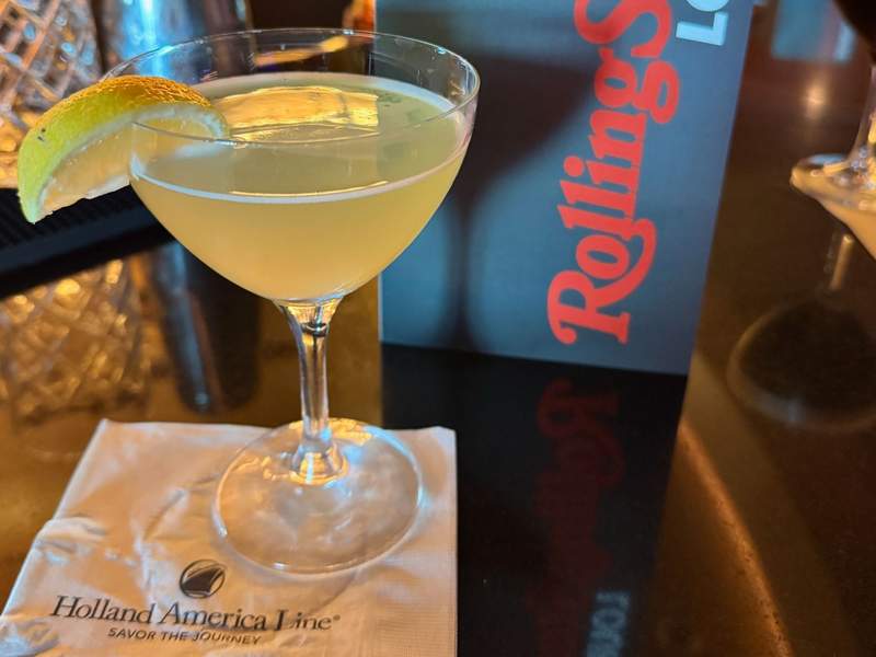 Light yellow cocktail with lemon quarter on the side on a Holland America Line napkin and Rolling Stone Lounge menu.