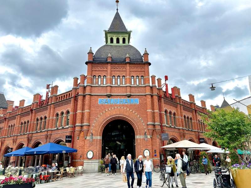 People gather in the square outside the red-brick Östermalms Saluhall, with its large arched entrance.