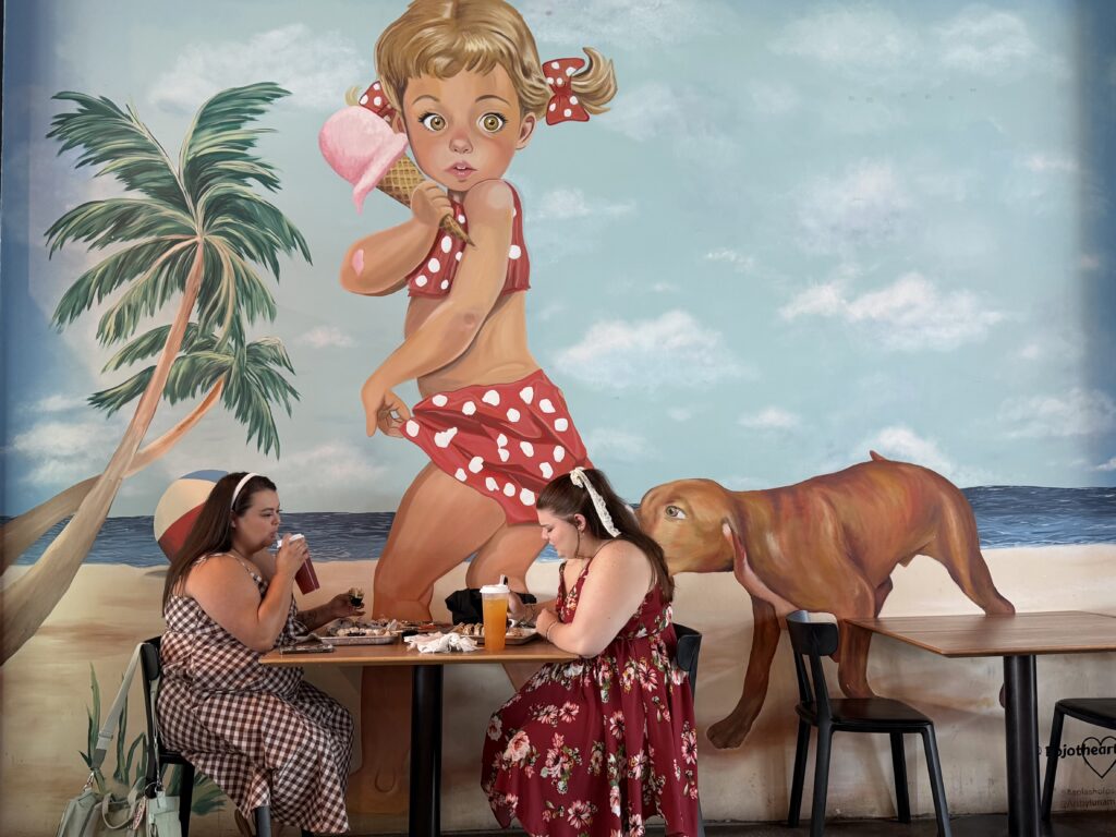 Two women eating at a table in front of a colorful mural of a girl with an ice cream and a dog in Tampa