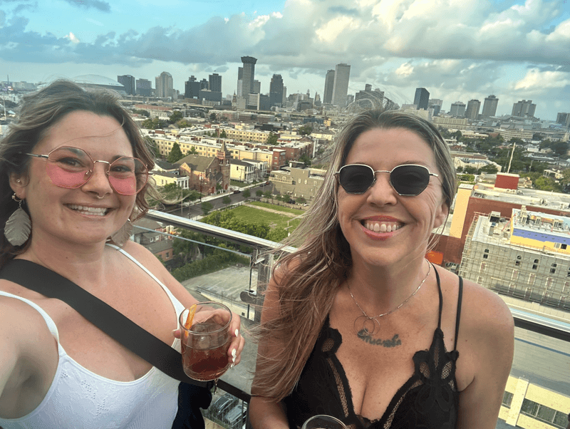 The writer and a friend smile at the camera while holding cocktails at the Hot Tin bar on the rooftop of the Pontchartrain Hotel in New Orleans.
