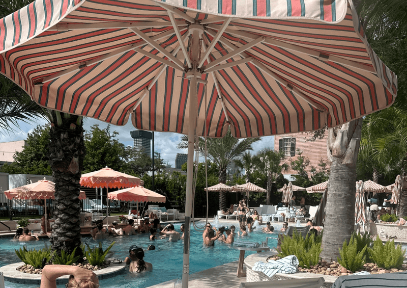 Resort style pool at the Garden District Hotel in New Orleans with striped umbrellas palm trees and guests swimming and lounging.