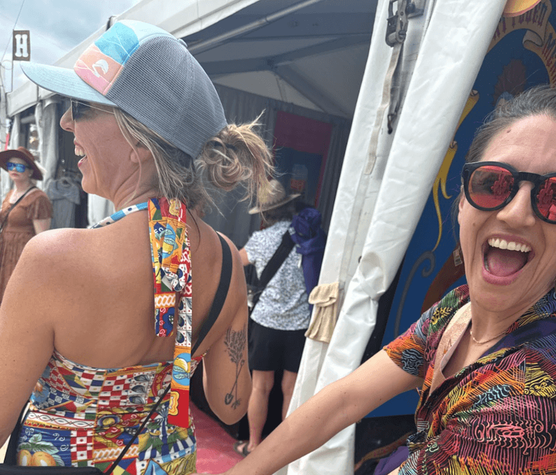 Two women laughing at Jazz Fest in New Orleans one wearing a colorful patterned swimsuit top and a light blue Sunday Afternoons trucker hat with sunglasses