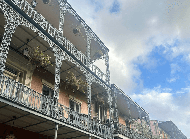 Looking up at ornate cast iron lace balconies on a multistory building in the French Quarter in New Orleans with hanging ferns and a cloudy blue sky.