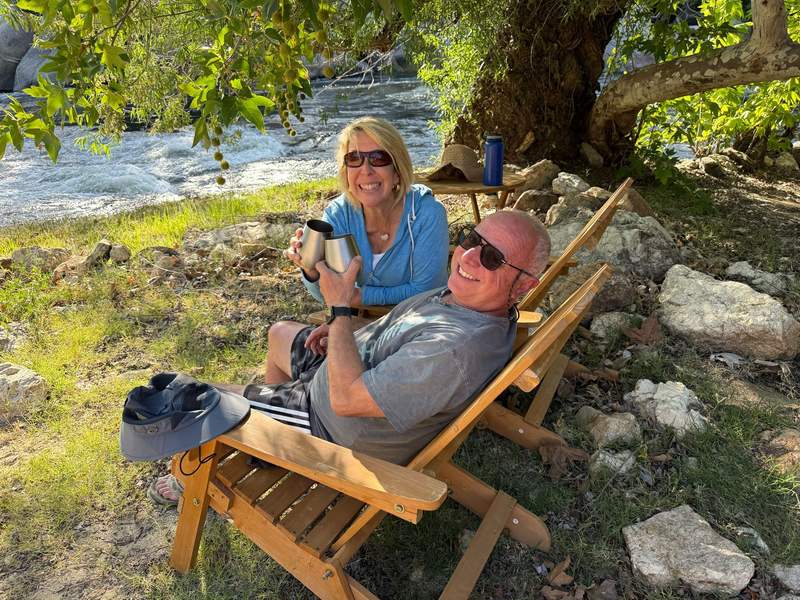 Author and husband relaxing in lawn chairs under a tree next to the river.