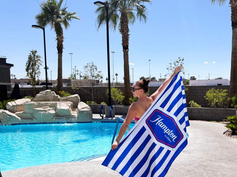 Woman holding up a blue and white Hampton by Hilton towel in front of a swimming pool.