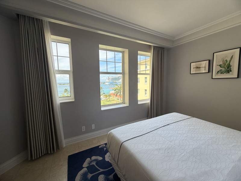 A bright guest room at the British Colonial Nassau in the Bahamas featuring a bed with a white coverlet and three large windows overlooking the ocean and cruise ship harbor.