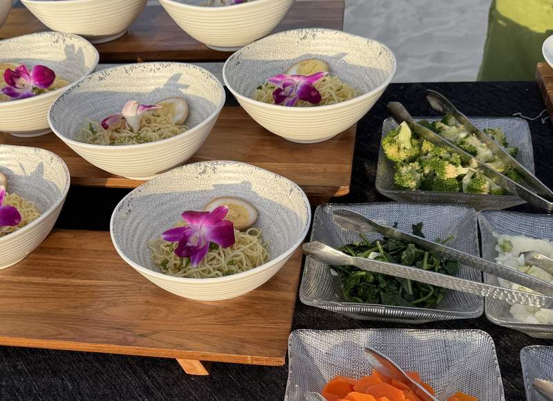 A resort buffet with bowls of ramen noodles topped with half a soft boiled egg and purple orchid alongside fresh vegetable toppings at the British Colonial Nassau.