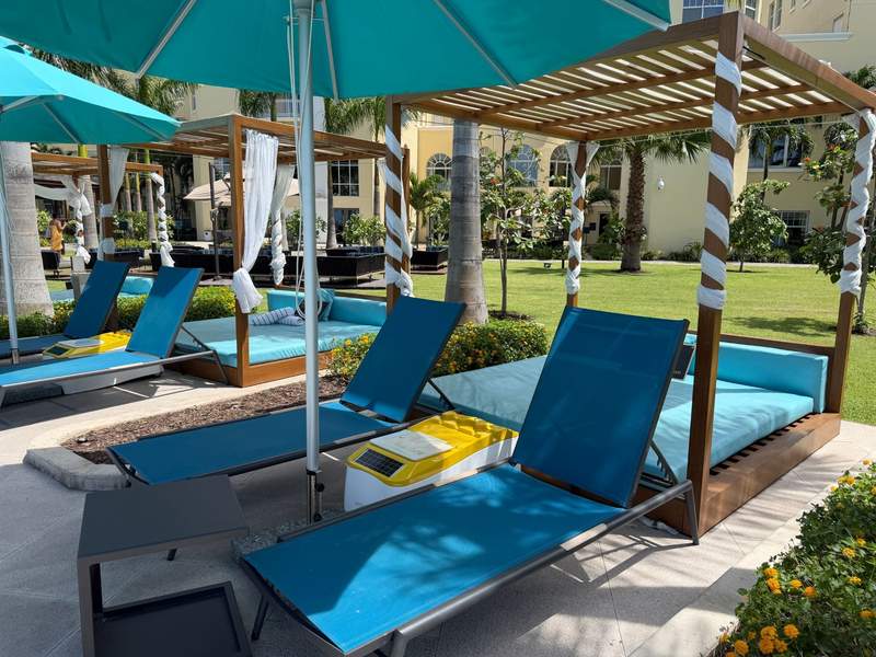 Pool patio at the British Colonial Nassau resort in the Bahamas, with bright teal lounge chairs wooden shade cabanas with draped white curtains and lush tropical landscaping.
