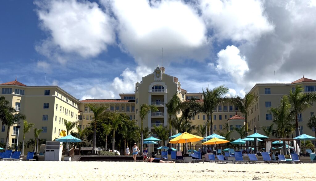 View from the sandy beach looking toward the pastel yellow facade of the British Colonial Nassau hotel, featuring palm trees and blue and yellow patio umbrellas.