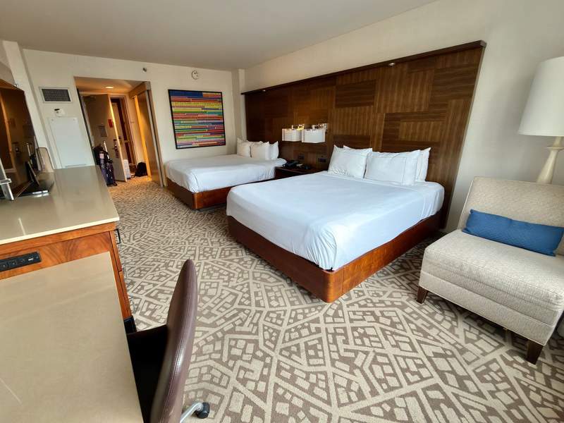 Two queen beds with white duvets and a white chair with blue pillow, patterned beige carpet.