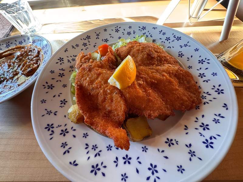 A crispy fried port schnitzel with a slice of lemon on top a bed of salad and roasted potatoes. 