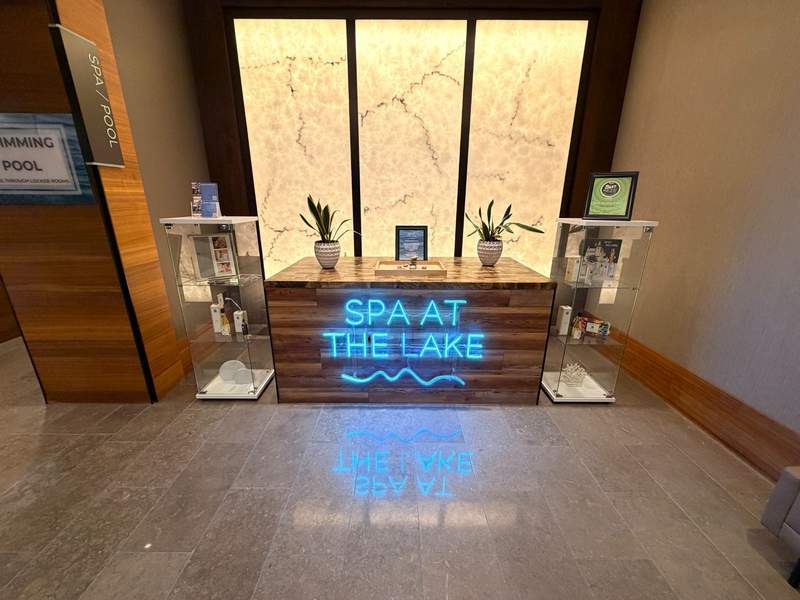 Entrance desk to the spa with a neon sign, Spa At The Lake and two glass kiosks with sample products for sale.