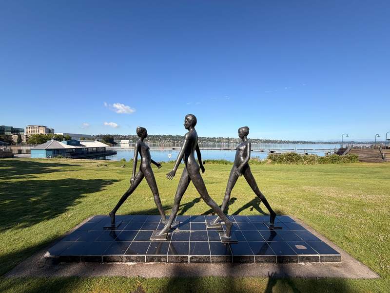 A bronze sculpture of two women and one man with elongated shapes walk on a granite slab with Lake Washington in the background.