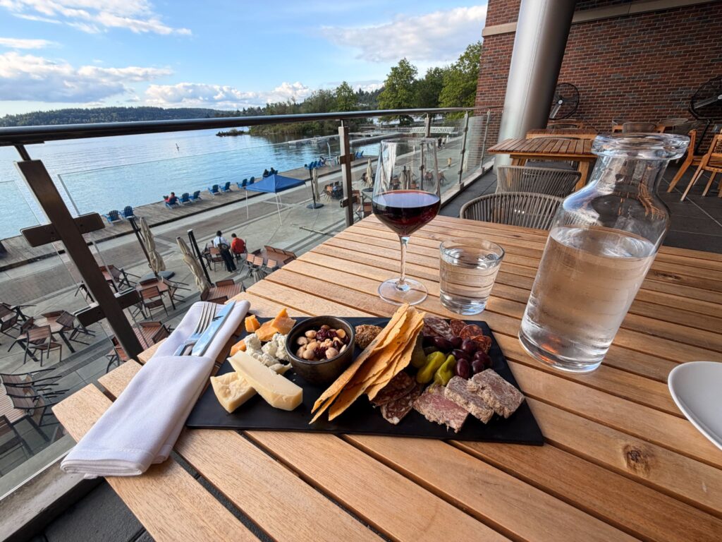 A charcuterie board on a black board with a white napkin and cutlery to the left. A carafe of water, a glass of wine and a glass of water at a table overlooking Lake Washington