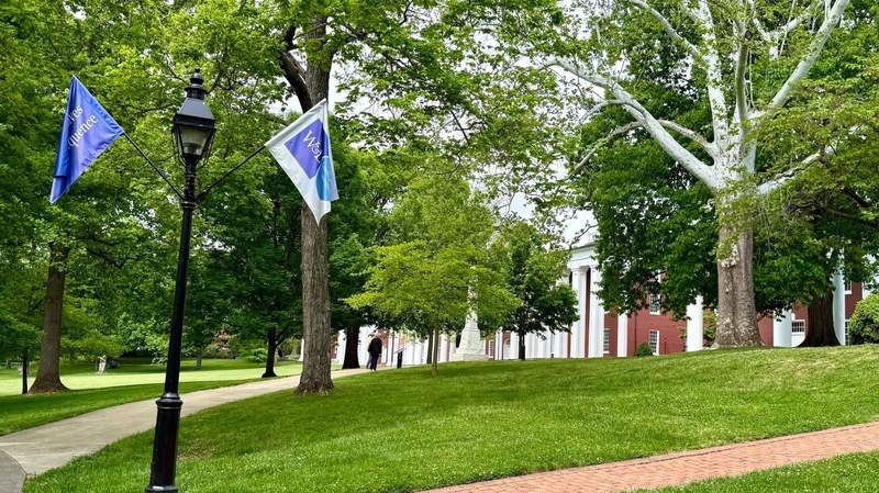 Green lawn flanked with pale brick sidewalks slopes uphill to a red brick two story building lined with white columns. Many leafy trees dot the lawn.