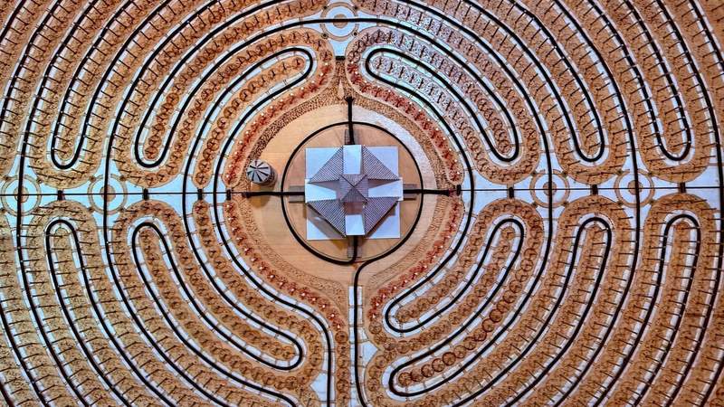 A painted labrynth hangs on the wall behind taps to serve cider. The colors are brown and sky blue. In the center is a picnic pavilion.