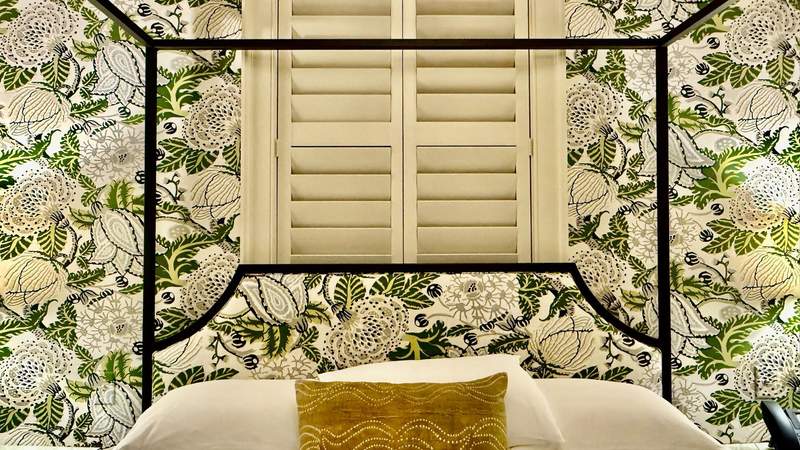 Closeup view ot a bed with matching wallpaper and headboard fabric. Swirls of green leaves and white flowers surround a window covered with off white shutters.