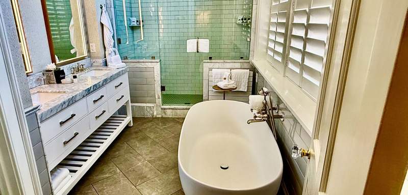 A high sided white porcelain tub sits across from a counter with two sinks, two framed mirrors and a slatted shelf for towels. The walk in shower at the end of the room fills the horizontal space.