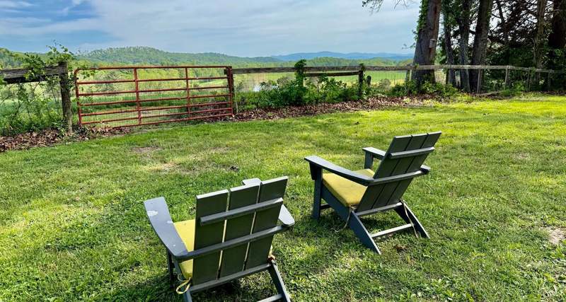 Two empty wooden arm chairs with slanted backs overlook blue sky filled with horizontal white clouds and a split rail fence.
