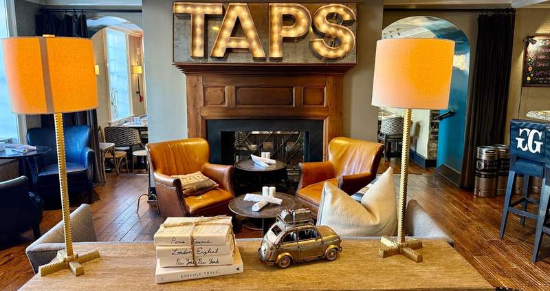 Four lighted capital letters spelling TAPS hang over a fireplace with two brown leather arm chairs in front. A wooden table holds two lamps four books and a toy car.