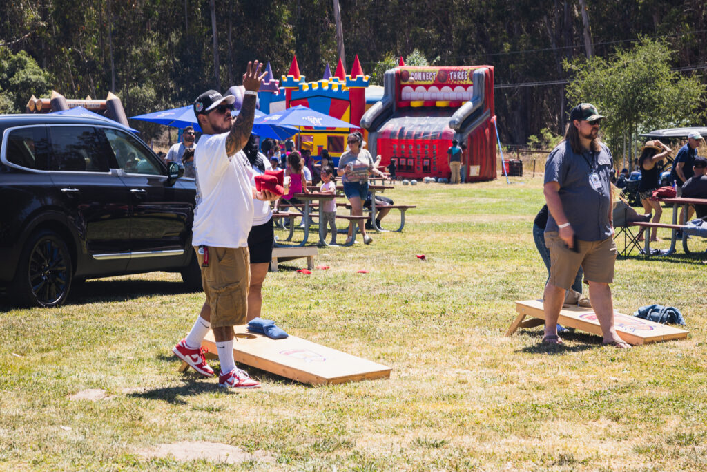 2 men playing corn hull with picnic tables and family-friendly bounce houses in the background
