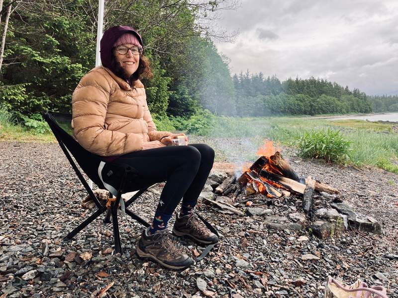 A woman relaxing alone , dressed warmly by a crackling fire, in a camp chair along a rocky Alaskan shoreline.
