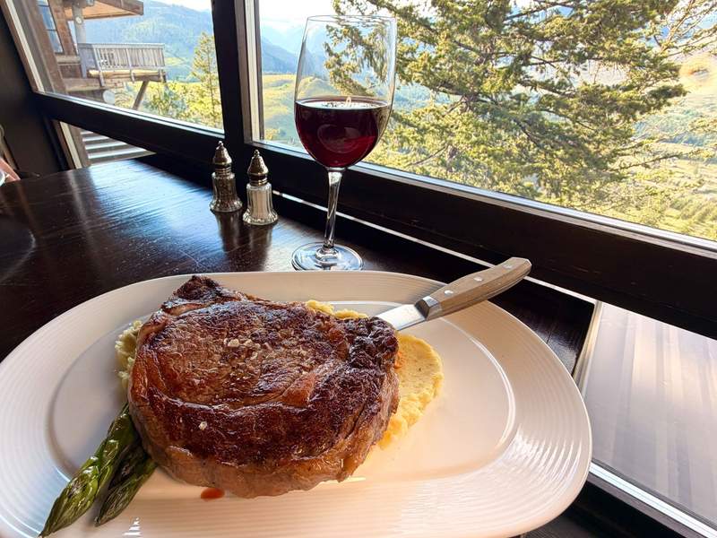 A large ribeye steak with local asparagus, polenta, and a knife, set beside red wine with scenic mountain views through floor-to-ceiling windows.