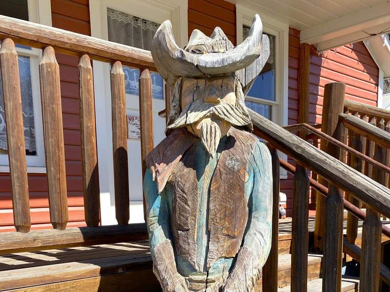 Life-size wooden carving of a western sheriff in a cowboy hat and leather vest, standing by wooden stairs beside a red building.