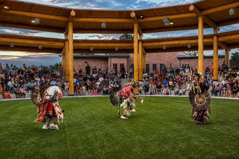 Native American dancers in colorful regalia performing for a seated crowd beneath the open-air performance arbor at Morning Star American Indian Village.
