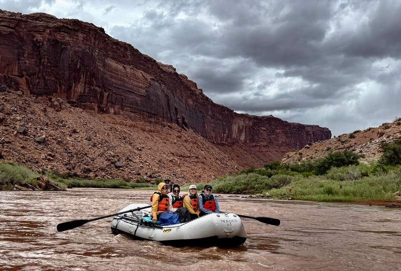 Horizontal view of a raft floating down the calm waters of the Colorado River surrounded by red canyon walls