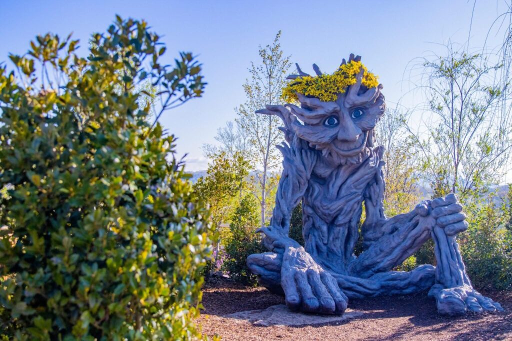 Large carved tree creature sculpture with a smiling face and yellow flowers on its head, sitting among landscaped greenery under a clear blue sky at Anakeesta in Gatlinburg, Tennessee.