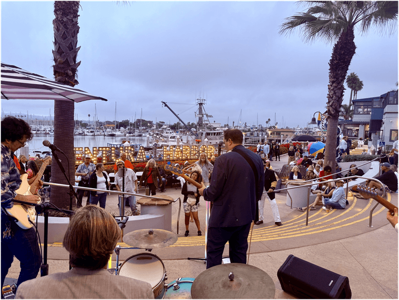 band performing on an outdoor stage near boats at Ventura Harbor with a crowd watching