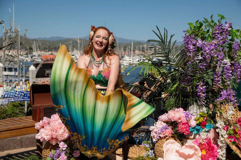 woman in a mermaid costume with a green and blue tail smiling surrounded by flowers and boats