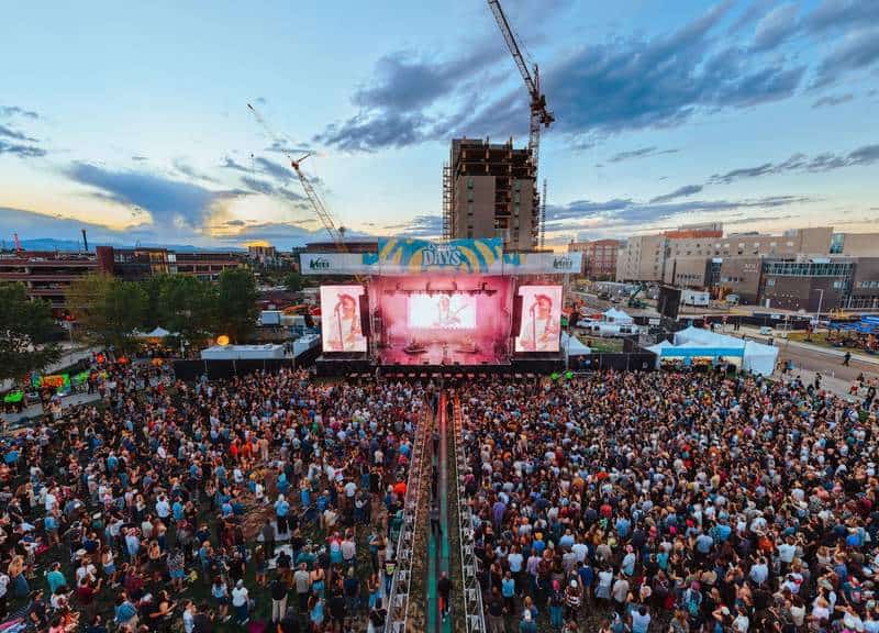 A crowd gathers near outdoor festival stages surrounded by greenery at Outside Days under clear blue skies in Denver.