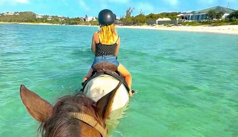 View from horseback of a girl riding a horse through turquoise ocean water in the Turks and Caicos.