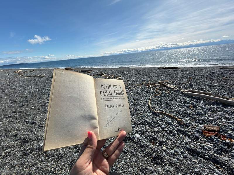 A hand holds an open, signed copy of Death on a Casual Friday by Sharon Duncan against a backdrop of the rocky shoreline and sparkling Salish Sea at South Beach on San Juan Island.