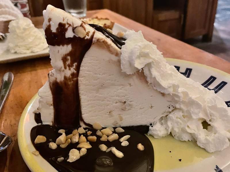 A towering slice of Duke’s Hula Pie with macadamia nut ice cream, glossy hot fudge, chopped macadamia nuts and clouds of whipped cream on a plate.