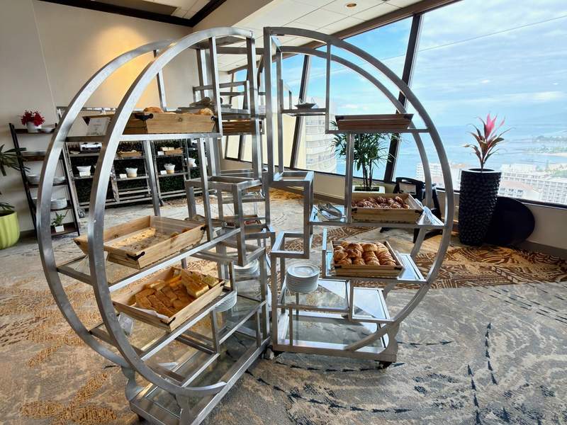 Golden croissants and pastries fill a sculptural breakfast display beside floor-to-ceiling windows overlooking blue ocean and Waikiki.