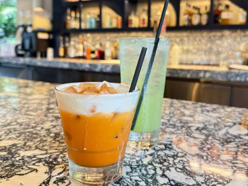 Two bright tropical cocktails one creamy orange and one pale green sit on a marble bar at Redfish in Waikiki.