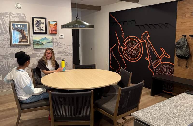 Two women sitting at a round wooden dining table inside a suite at the Eddie Bauer Adventure Club in Moab with desert artwork and a bike storage feature wall.