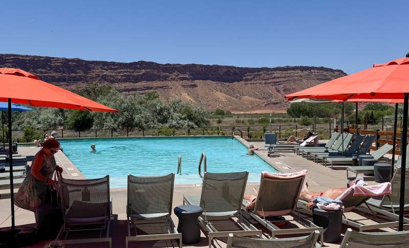 Outdoor heated swimming pool surrounded by lounge chairs and red umbrellas with steep red rock cliffs in the background.