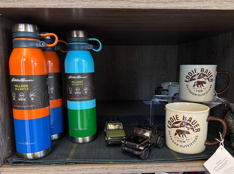 Colorful Eddie Bauer Hillside water bottles and outdoor themed ceramic mugs displayed on wooden shelving.