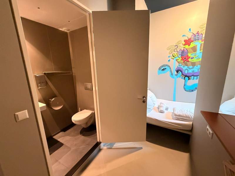 A private room in an Amsterdam hostel features a single bed with fresh linens, colorful wall art and an adjoining private bathroom with a toilet and shower.